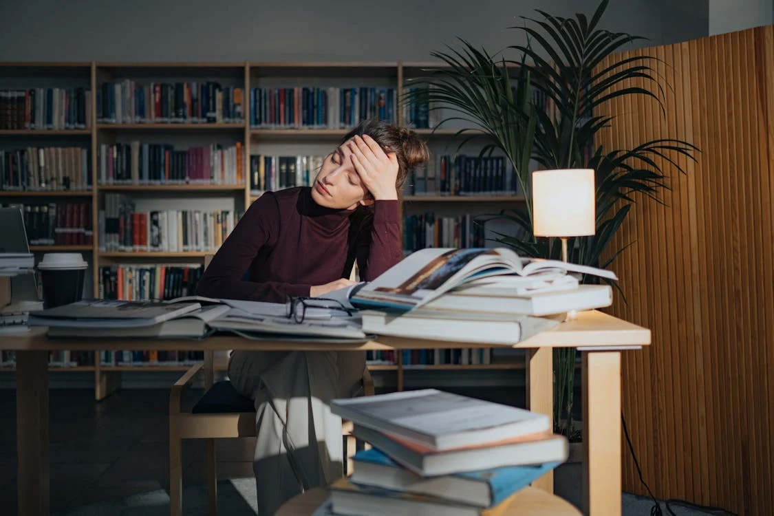 A stressed woman sitting at a library desk with multiple open books and papers, holding her forehead in frustration. The background features bookshelves, a lamp, and a potted plant. The image illustrates the challenges of studying for the California QME Competency Examination and highlights the importance of effective preparation strategies.