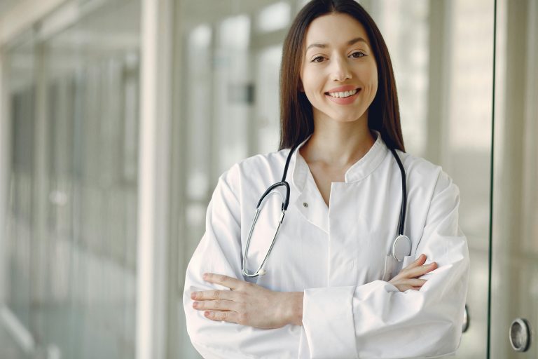 A smiling female doctor wearing a white coat and a stethoscope, standing in a bright hallway, conveying confidence and professionalism in a medical setting.