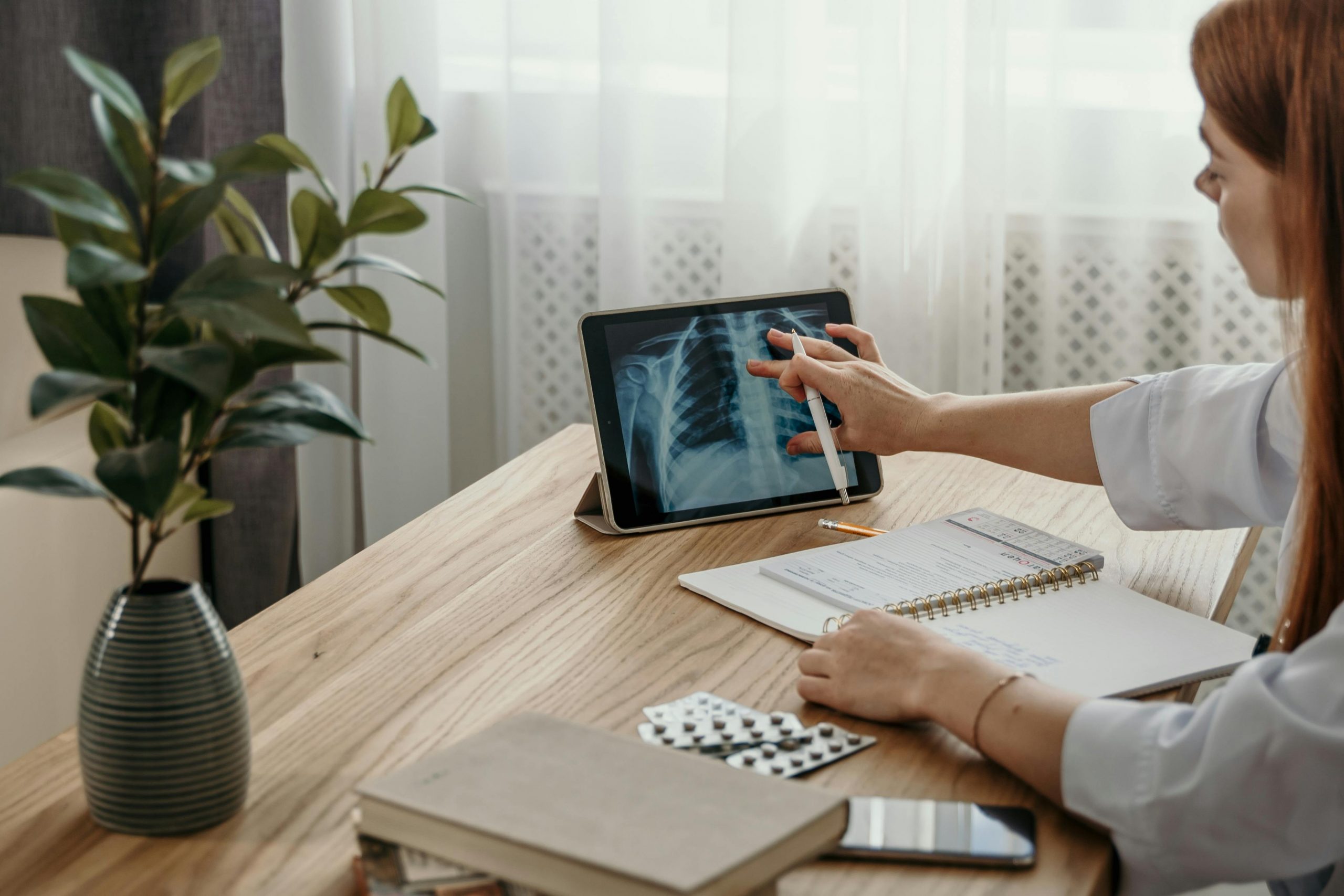 A female doctor sitting at a desk, reviewing an X-ray displayed on a tablet while taking notes in a notebook, representing study and preparation for a medical exam.