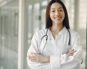 A smiling female doctor wearing a white coat and a stethoscope, standing in a bright hallway, conveying confidence and professionalism in a medical setting.